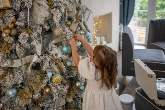 A girl in a white dress decorates a Christmas tree with her own hands, Christmas holidays at home