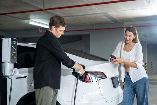 Young couple travel with EV electric car to shopping center parking lot charging in downtown city showing urban sustainability lifestyle by green clean rechargeable energy of electric vehicle innards