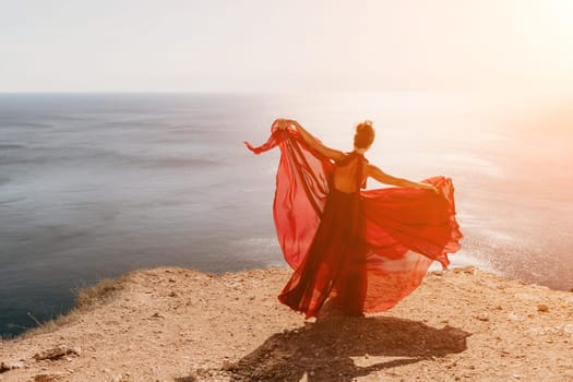 Side view a Young beautiful sensual woman in a red long dress posing on a rock high above the sea during sunrise. Girl on the nature on blue sky background. Fashion photo.