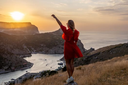 Happy woman standing with her back on the sunset in nature in summer with open hands posing with mountains on sunset, silhouette. Woman in the mountains red dress, eco friendly, summer landscape active rest.