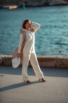 Happy blonde woman in a white suit and hat posing at the camera against the backdrop of the sea.