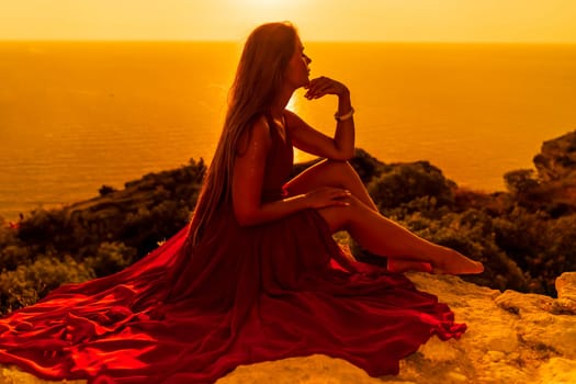 Woman sunset sea red dress, side view a happy beautiful sensual woman in a red long dress posing on a rock high above the sea on sunset