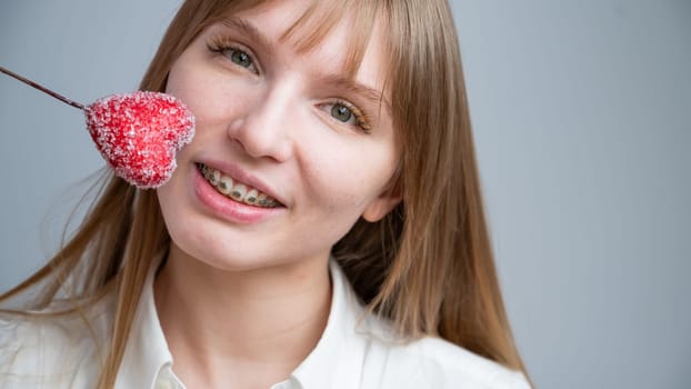 Cute woman with braces on her teeth holds a candy in the form of a heart on white background. Copy space.