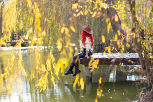 Autumn lake woman. In autumn, she sits by the pond on a wooden pier and admires nature with red hair and a hat. Tourism concept, weekend outside the city