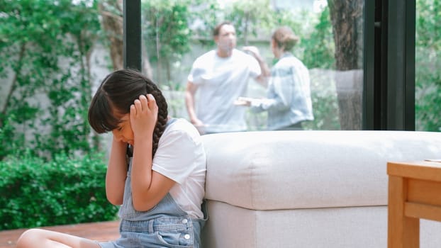 Stressed and unhappy young girl huddle in corner, cover her ears blocking sound of her parent arguing in background. Domestic violence at home and traumatic childhood develop to depression. Synchronos