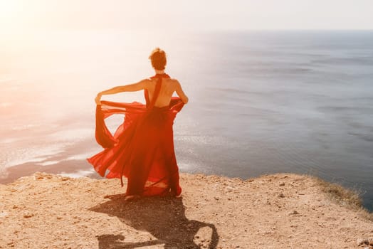 Side view a Young beautiful sensual woman in a red long dress posing on a rock high above the sea during sunrise. Girl on the nature on blue sky background. Fashion photo.