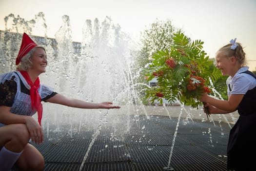 Young and adult schoolgirl on September with flowers having fun near water of fontain. Generations of schoolchildren, pioneer of USSR and October girl in modern uniform of Russia. Mom and daughter