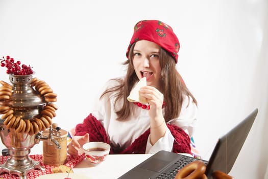 Girl in a red colored scarf, white shirt and shawl near a table with a samovar, bagels drinks tea for the Easter holiday and Masolennitsa. A freelancer or blogger with a laptop is resting and working