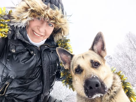 Adult girl with shepherd dog in tinsel taking selfie in winter day. Middle aged woman and big shepherd dog on cold nature. Friendship, love, fun, hugs