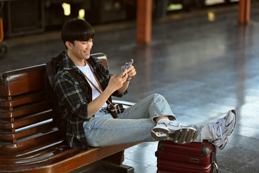 Happy Asian male traveler with suitcase using mobile phone while waiting train. Travel and vacations concept
