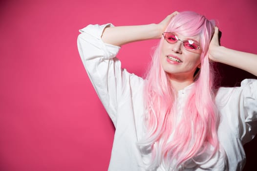 Close-up portrait of a young woman with braces in a pink wig and sunglasses on a pink background. Copy space