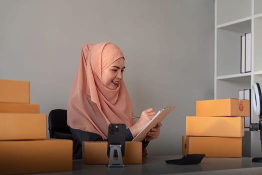 Muslim women selling online at home with box. Selling online with box to accept order from customer. SME business idea. Parcel delivery. muslim woman working smartphone and laptop at home.