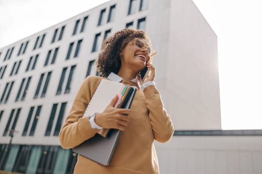 Smiling woman manager is talking phone while standing with laptop on modern building background