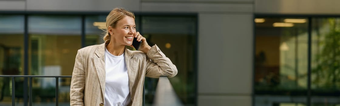 Smiling female manager talking phone standing on modern office building background