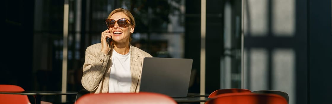 Businesswoman in eyeglasses talking on phone with client while working on laptop in office