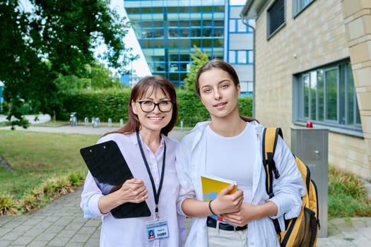 Teenage girl high school student with backpack talking to female teacher, mentor, coach, standing outdoors on educational building background. Adolescence, education, knowledge, communication