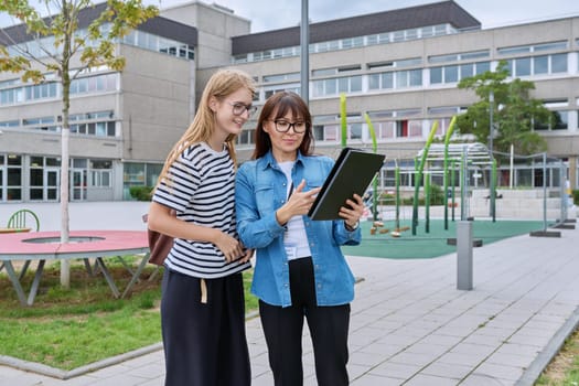 Talking female teacher and teenage high schoolgirl outdoor, school building background. Meeting communication student girl with backpack and mentor counselor. Education, adolescence, learning concept