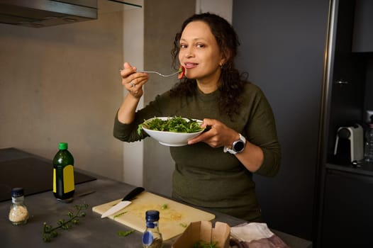 Beautiful multi ethnic young woman standing at kitchen table, holding a fork with salad near her face, smiles cutely looking at camera. Happy conscious female eating healthy meal. People. Lifestyle