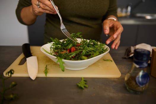 Details on the hands of a woman holding fork and a white bowl of fresh healthy salad with organic vegetables and greens, while preparing healthy dieting meal for dinner in a cozy home kitchen interior