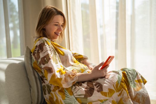 Pregnant joyful woman checks her smartphone relaxing on the sofa.