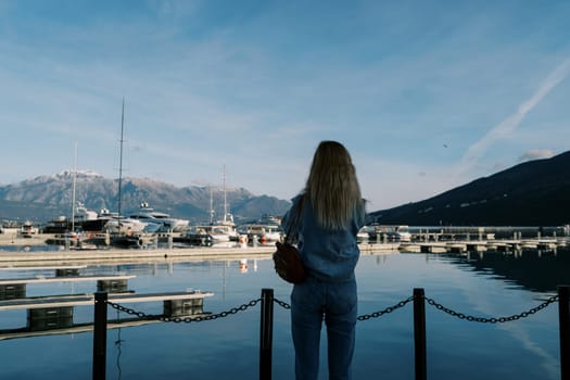 Woman stands on a pier by the sea and looks at the moored yachts. Back view. High quality photo