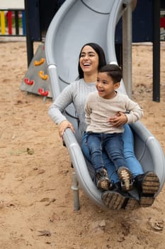 Smiling latin mom having fun playing on the slide together with her son in an outdoor playground.