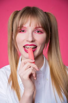 Portrait of a young woman with braces and bright makeup chewing gum on a pink background. Vertical photo