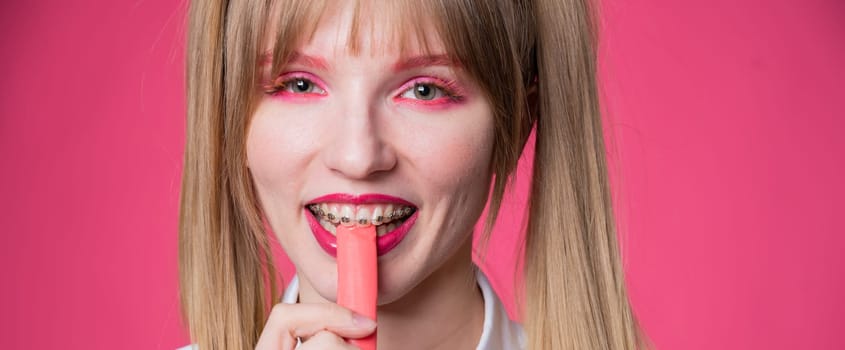 Portrait of a young woman with braces and bright makeup chewing gum on a pink background