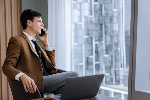 Closeup of handsome businessman making phone call with manager while sitting near window with skyscraper view. Executive manager talking working by using phone and laptop. Look aside. Ornamented.