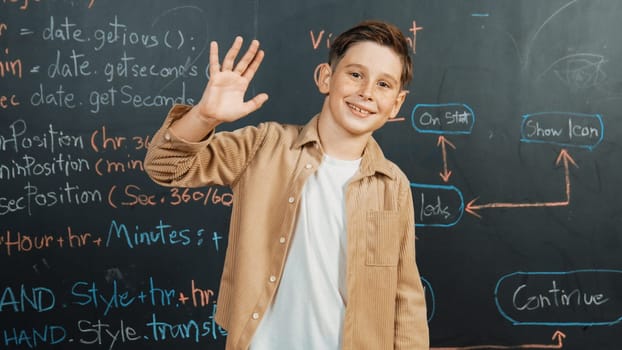 Panorama shot of happy caucasian boy smiling and waving hand at blackboard with engineering code or prompt written. Smart child looking at camera and greeting while study in STEM classroom. Erudition.