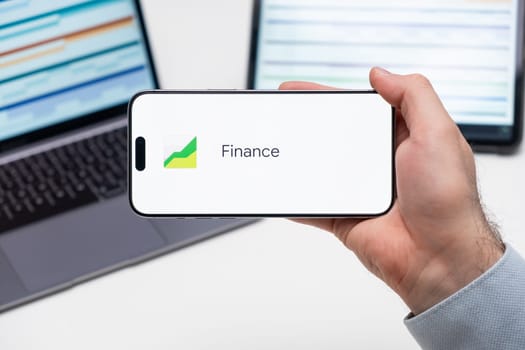 Finance logo on the screen of smart phone in mans hand, laptop and tablet are on the table in the background, December 2023, Prague, Czech Republic.