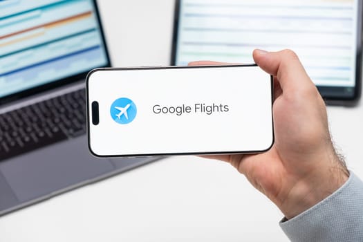 Google Flights application logo on the screen of smart phone in mans hand, laptop and tablet are on the table in the background, December 2023, Prague, Czech Republic.