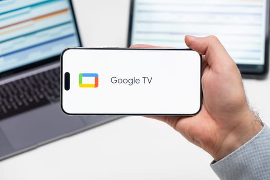 Google TV logo of app on the screen of mobile phone held by man in front of the laptop and tablet, December 2023, Prague, Czech Republic