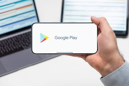 Google play logo of app on the screen of mobile phone held by man in front of the laptop and tablet, December 2023, Prague, Czech Republic