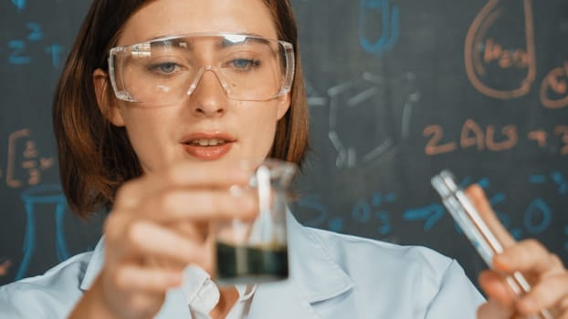 Closeup of young beautiful teacher focus on mixing chemical liquid at laboratory. Skilled scientist doing an experiment by inspect colored solution while standing in front of blackboard. Erudition.