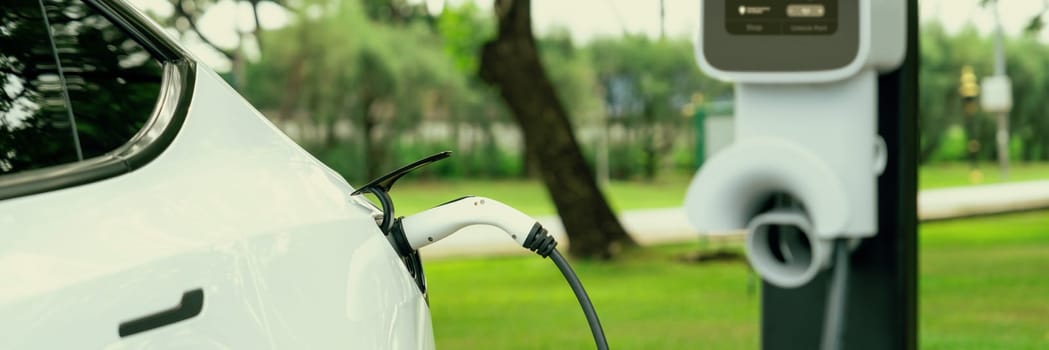 EV electric vehicle recharging battery from EV charging station in outdoor green city park scenic. Natural protection with eco friendly EV car travel. Panorama Banner Exalt