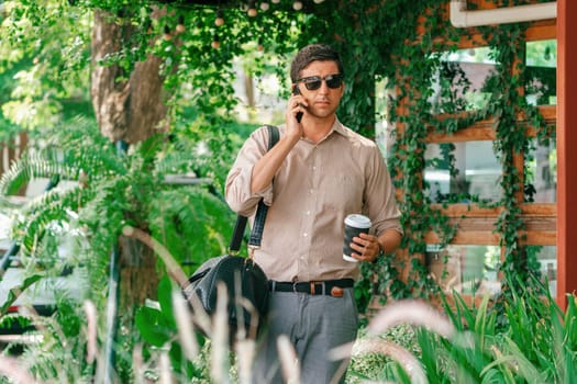 Young handsome man walk out of coffee shop with cafe garden, holding a coffee cup and a shopping bag while talking on the phone. Modern happy carefree with sunglasses lifestyle. Expedient