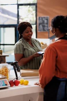 An African American vendor smiles as a customer uses contactless payment at a bio food market. Freshly harvested, organic produce is available for sale.