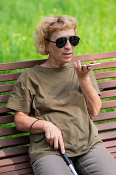An elderly blind woman sits on a bench in the park and talks on a smartphone