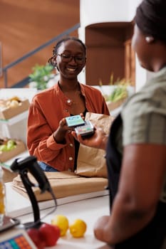 Female customer holding and using her mobile to device to make nfc payment for her sustainable goods. Young vendor holds out pos machine for client to pay with cell phone at loce eco friendly store.