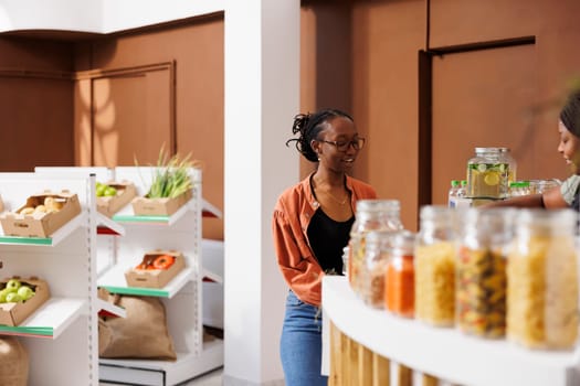 Portrait of african american female shopper standing at checkout counter, speaking with shopkeeper. Young lady with glasses waiting for her sustainable organic packaged products.