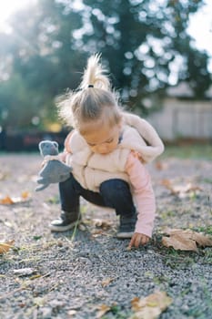 Little girl with a soft toy squats down on a gravel path and touches a pebble with her finger. High quality photo
