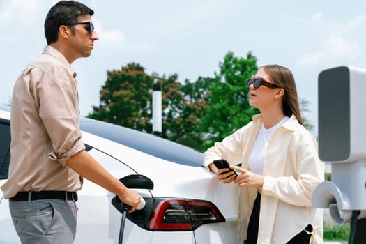 Young couple use smartphone to pay for electricity at public EV car charging station green city park. Modern environmental and sustainable urban lifestyle with EV vehicle. Expedient