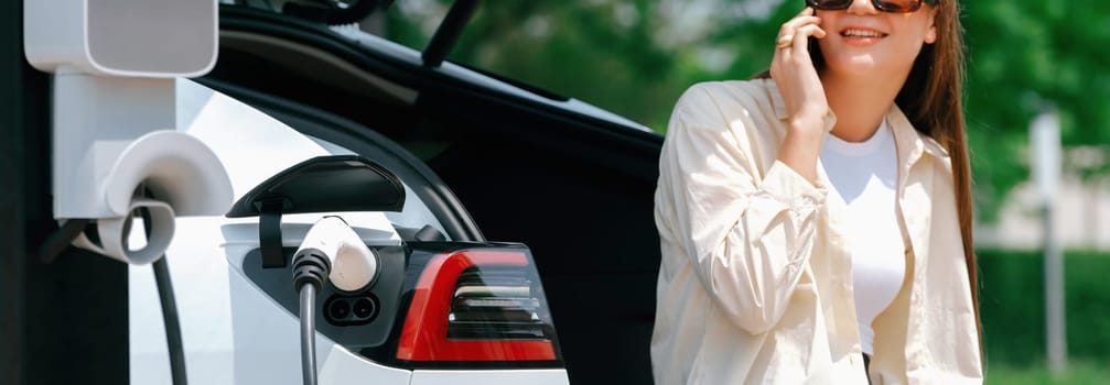 Young woman recharge EV electric vehicle at green city park while talking on phone. Sustainable urban lifestyle for environment friendly EV car with battery charging station. Panorama Expedient