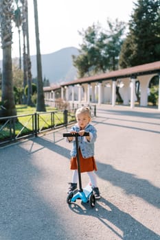 Little girl stands near a scooter holding the steering wheel on the road in a green park. High quality photo