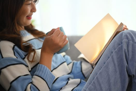 Happy young woman in warm sweater drinking hot herbal tea and reading book at home.