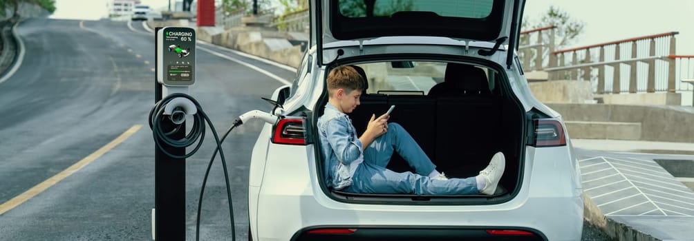 Little boy sitting on car trunk, using smartphone while recharging eco car from EV charging station. EV car road trip travel as alternative vehicle using sustainable energy concept.Panorama Perpetual