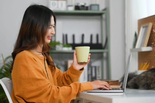 Side view of beautiful young woman holding coffee cup and reading email on laptop at home.