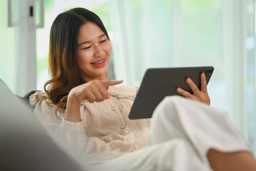 Shot of a happy young Asian woman using digital tablet while relaxing at home.