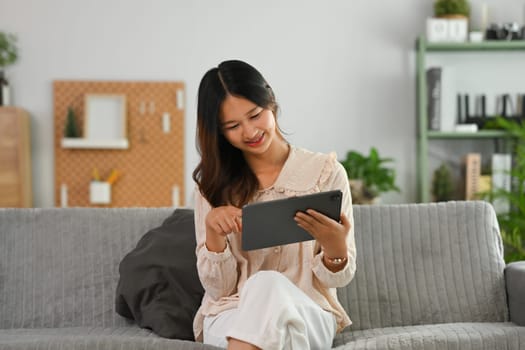 Smiling young woman in casual clothes using digital tablet on couch at cozy living room.